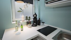 Une cuisine moderne avec un plan de travail blanc, une machine à café noire, un vase avec des fleurs blanches et une fenêtre avec des stores.