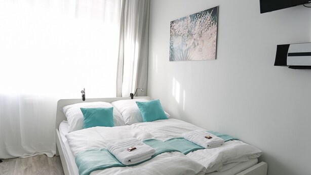 Un lit bien fait, avec une literie blanche et turquoise, une fenêtre ornée de rideaux légers et un climatiseur fixé au mur.