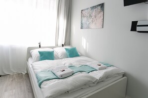 A neatly made bed with white and teal bedding, a window with sheer curtains, and a wall-mounted air conditioner. - Micro Appartments Katz (Nuremberg)
