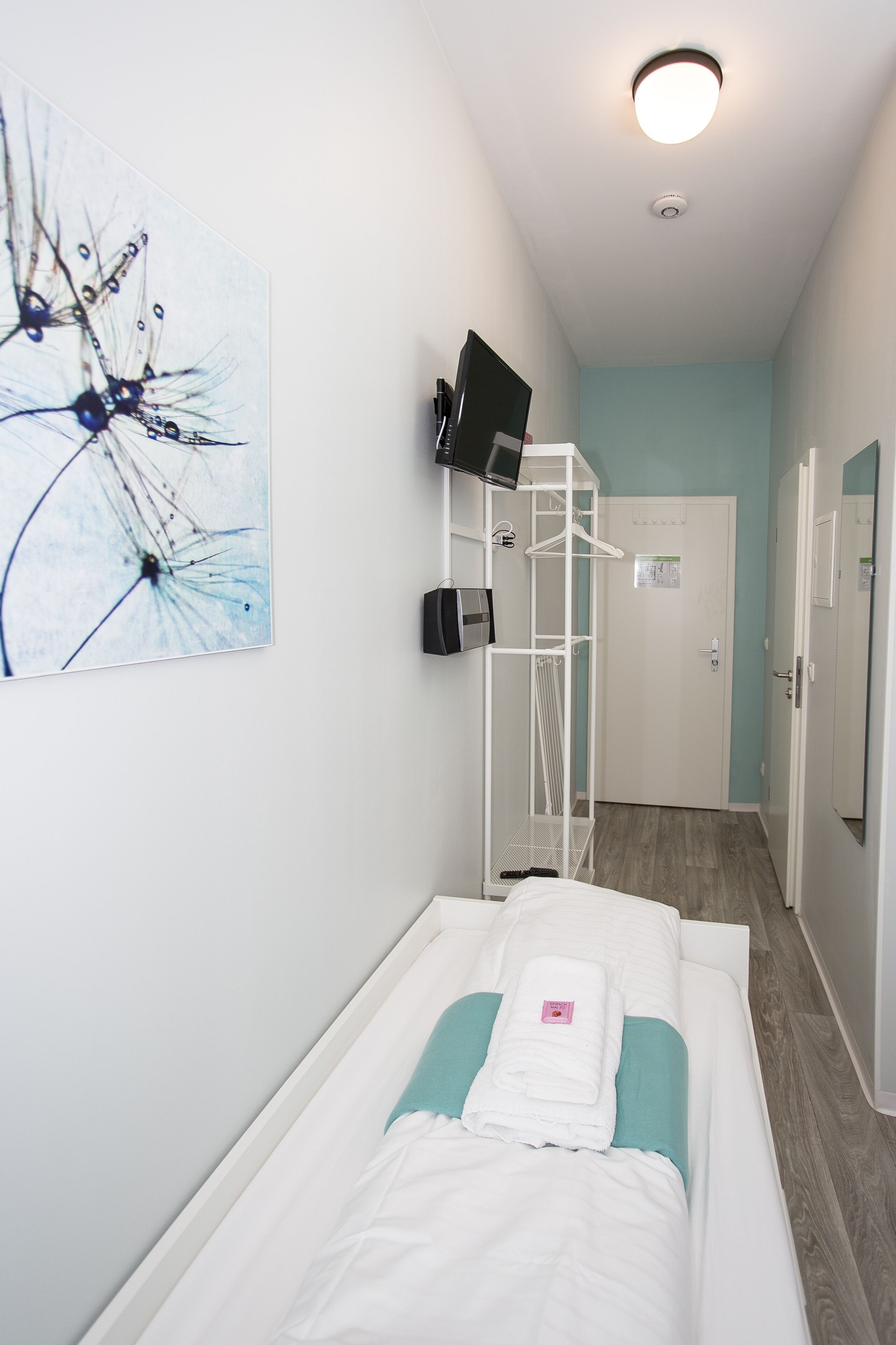 Habitación de hotel con una cama individual, televisor montado en la pared, una pizarra y una puerta que da a otra habitación.