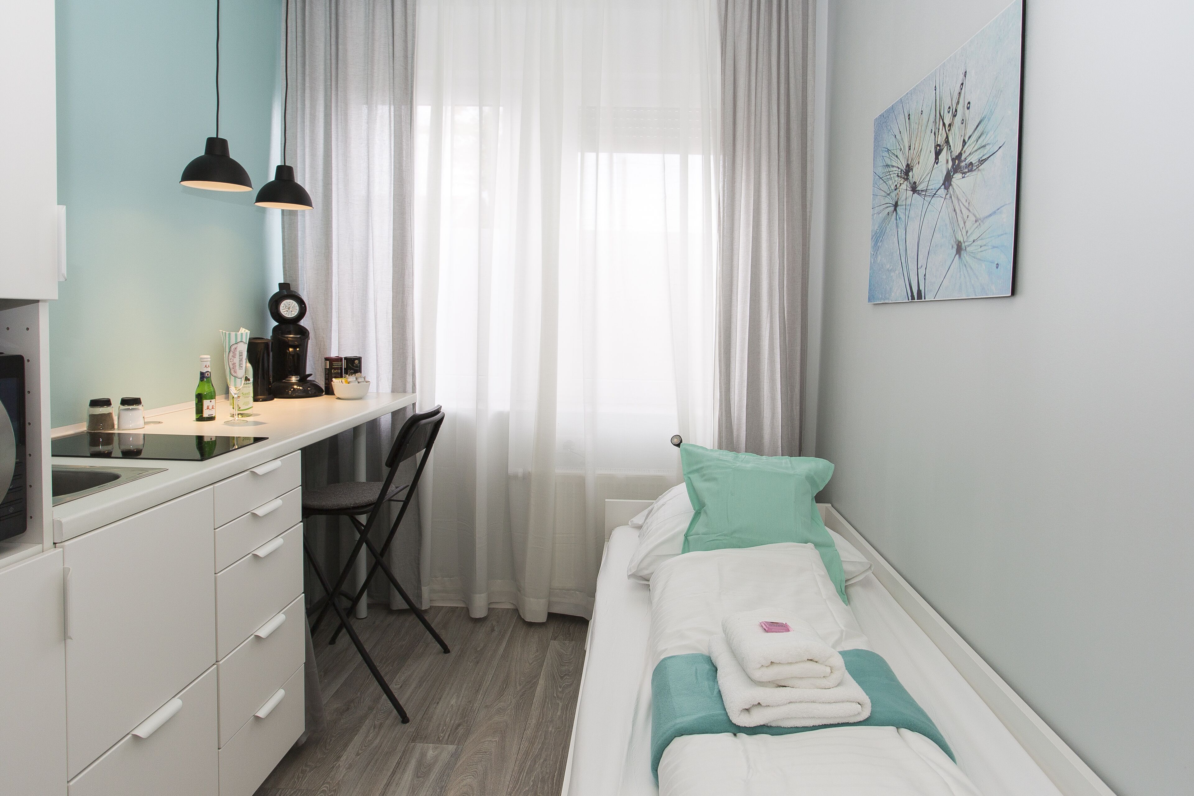 Una habitación de hotel compacta con kitchenette, una cama individual y una almohada verde azulado, y un cuadro en la pared.
