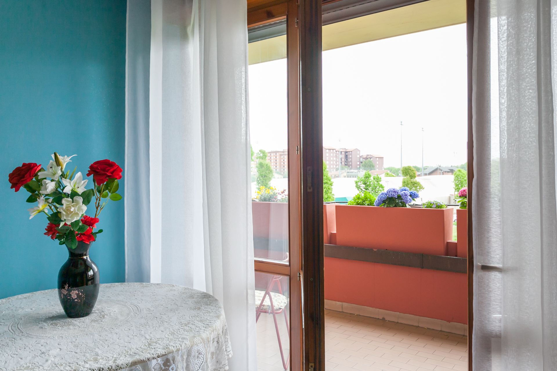 Una habitación con vistas a un balcón, una mesa con un jarrón de flores y una ventana con cortinas.