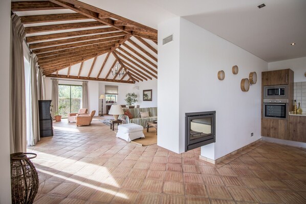 Un vaste espace de vie doté d’une cheminée, de poutres apparentes en bois au plafond et d’une cuisine équipée d’appareils électroménagers modernes.