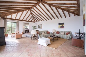 Un salon spacieux avec un plafond haut, des poutres apparentes, un ensemble de canapés, une table basse et un téléviseur.