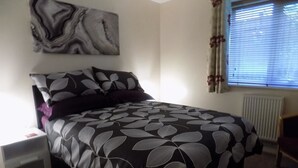 A neatly made bed with a leaf-patterned bedspread, a nightstand with a lamp, a wall-mounted abstract painting, and a window with blinds. - A Home From Home 3 (Plymouth)