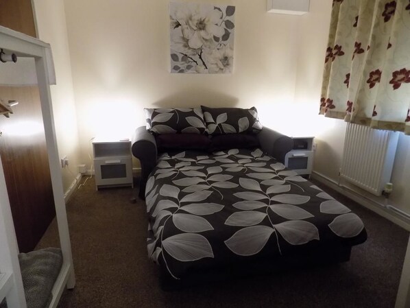 Quarto com cama, mesas de cabeceira, luminária na parede, radiador e cortina florida.