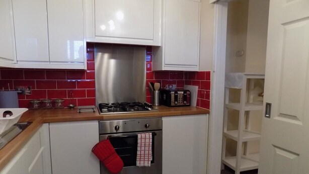 A kitchen with white cabinets, a red tiled backsplash, a stainless steel range hood, and a red oven mitt hanging on the oven door. - A Home From Home 3 (Plymouth)