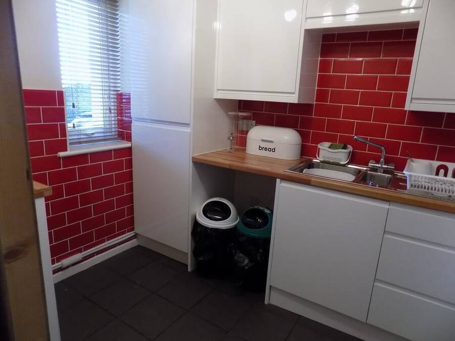 A kitchen with red tiled walls, white cabinets, a bread maker, and two trash bins.