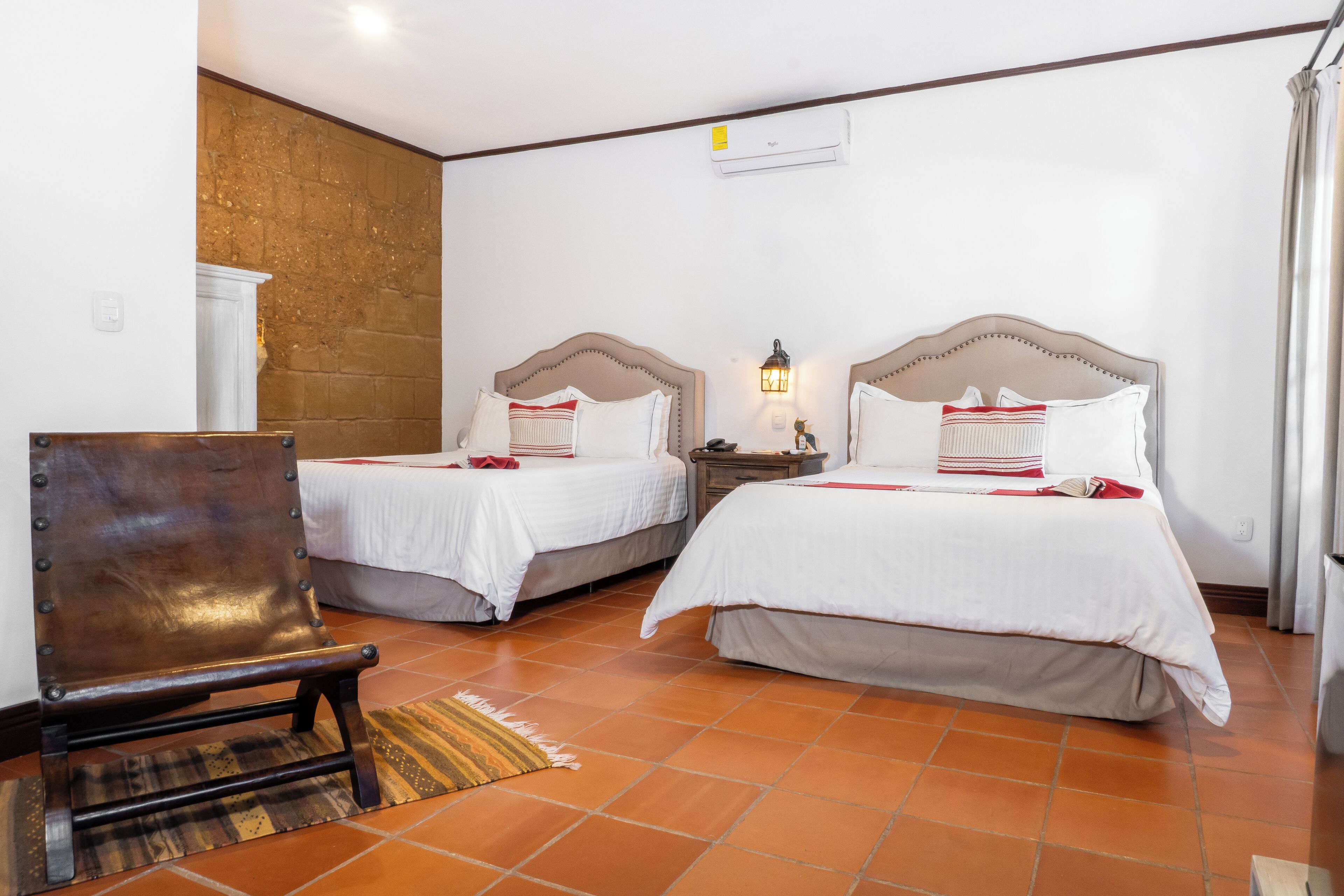 Une chambre d’hôtel avec deux lits, une chaise en bois, une table de chevet avec une lampe et un sol carrelé.