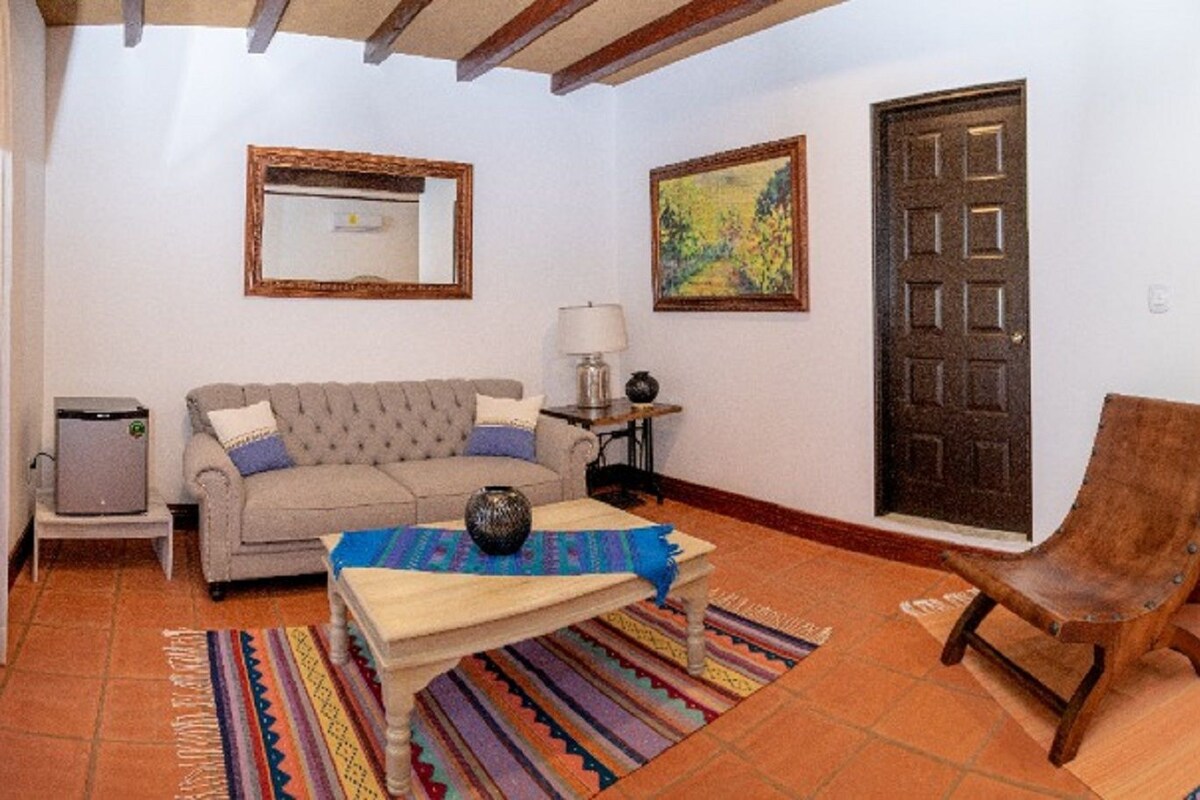 A hotel room with a sofa, two beds, a chair, a small table, a lamp, a mirror, and a door.