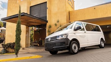 Una furgoneta Volkswagen blanca estacionada frente a un edificio, con una banquina amarilla, un árbol pequeño y cielo despejado.