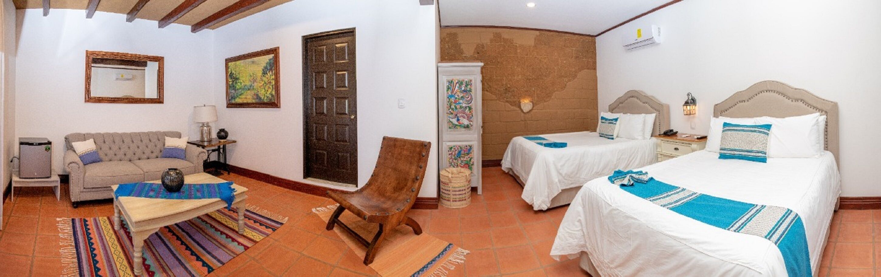 Habitación de hotel con sofá, dos camas, una silla, una mesita, una lámpara, un espejo y una puerta.