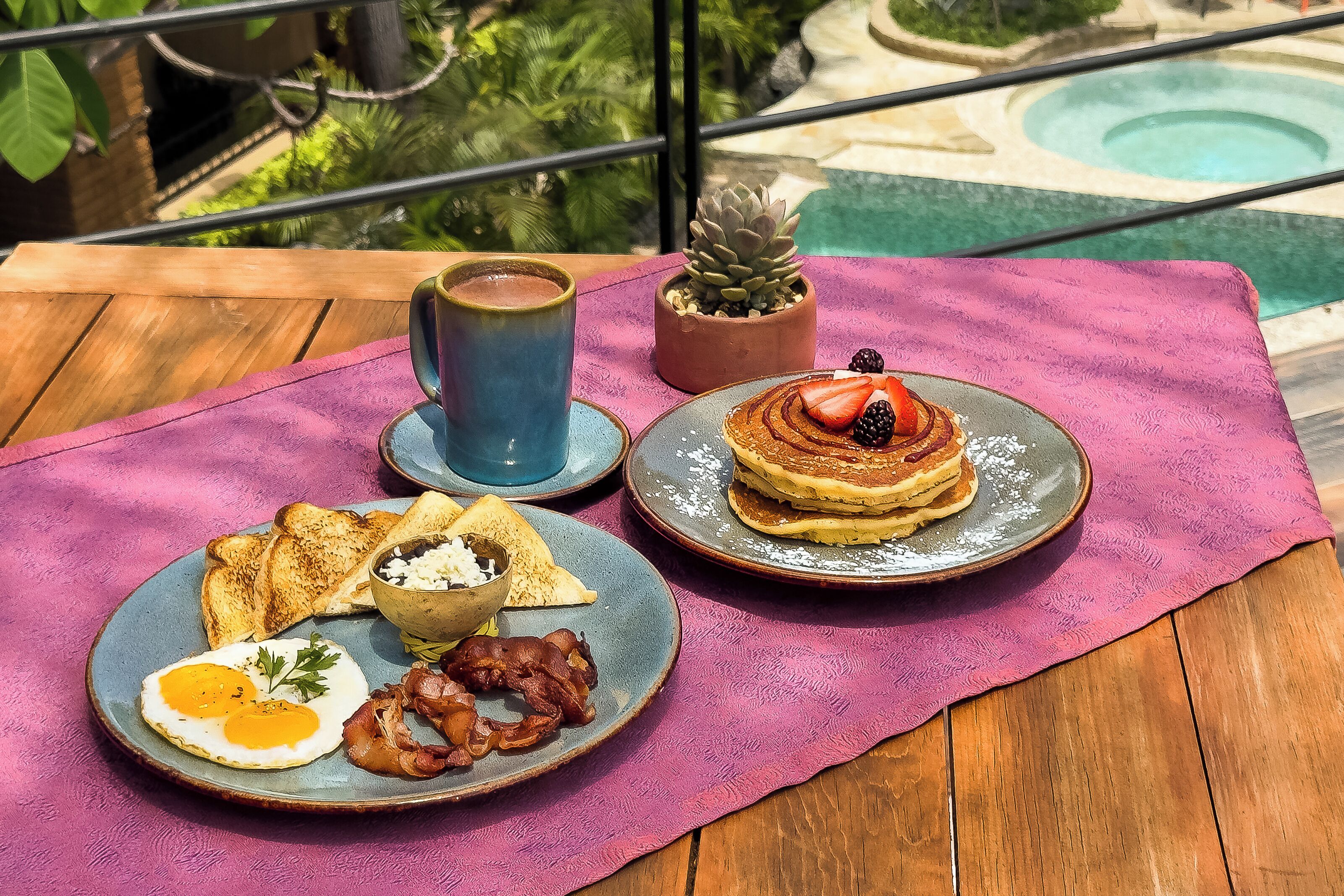 Disposição para o pequeno-almoço numa mesa de madeira com toalha roxa, incluindo torradas, ovos, bacon, panquecas com frutos vermelhos e uma chávena de café.