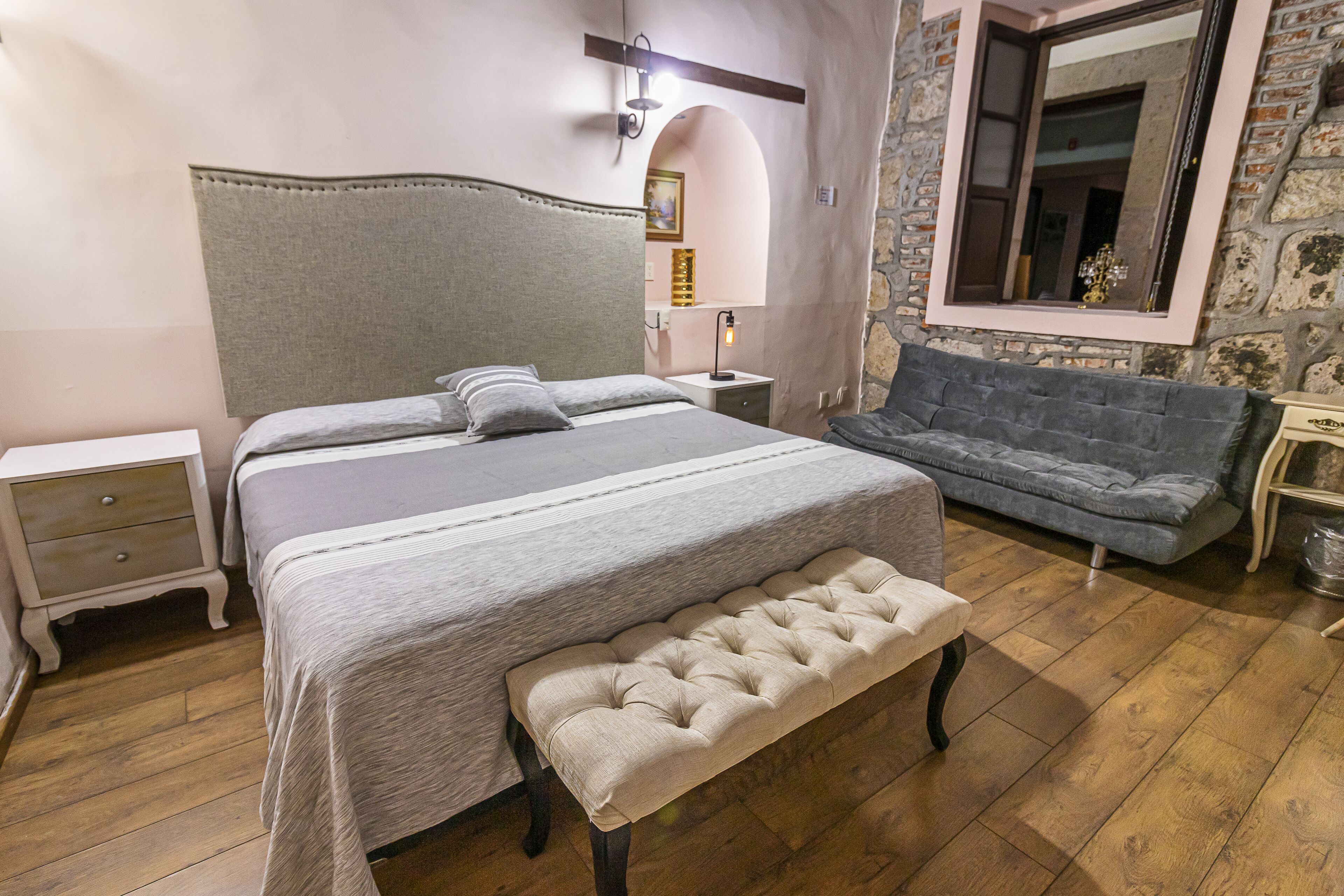 Un dormitorio con cama, banco, mesita de noche y una pared de piedra con espejo.