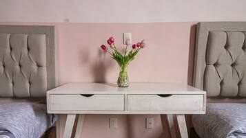 Ein Schlafzimmer mit weißem Schminktisch, zwei gesteppten Kopfteilen, einer rosa Wand und einer Vase mit Blumen auf dem Schminktisch.