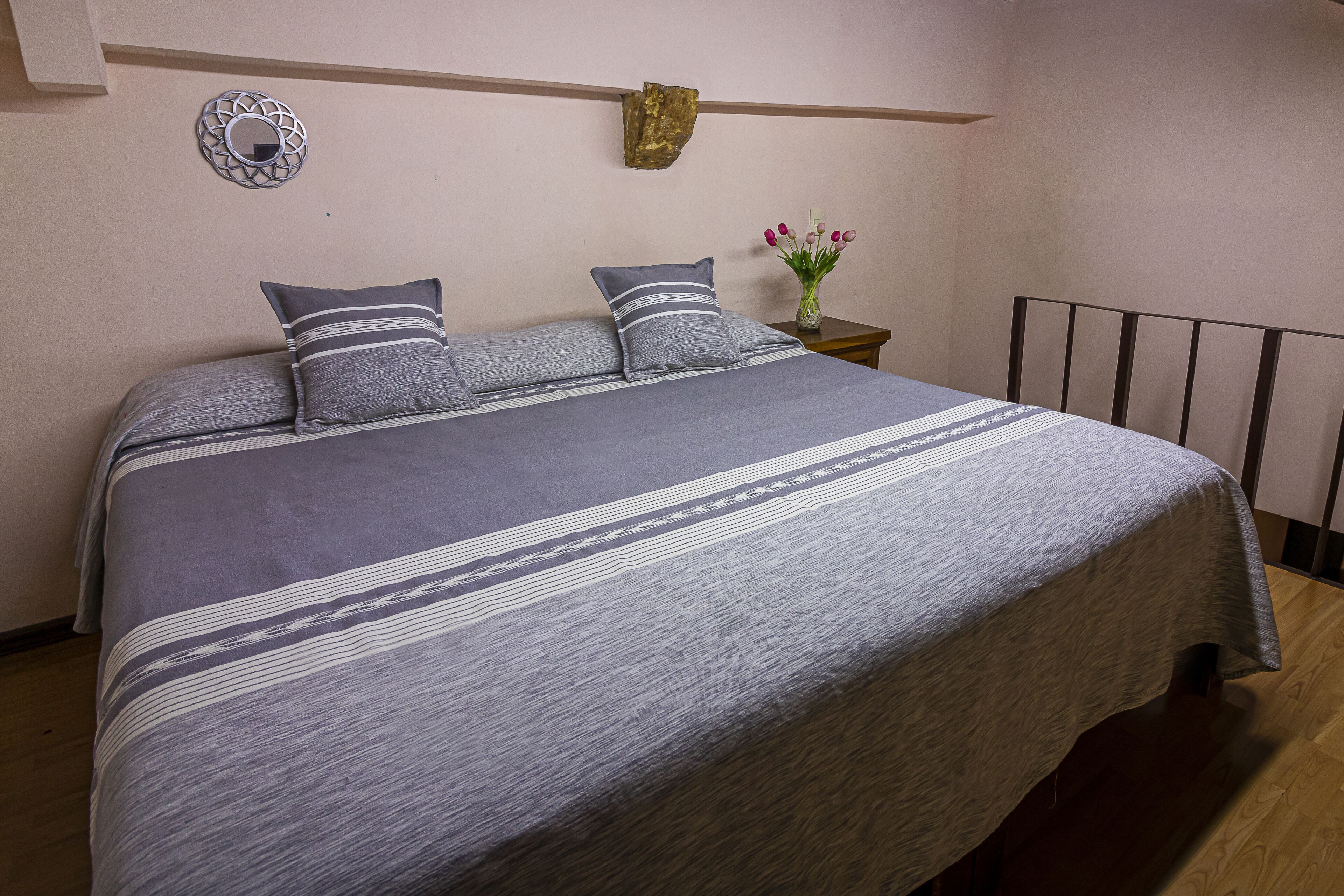 Een bed met een grijs-witte streepdeken, een klein nachtkastje met een bloemenvaas, en een spiegel aan de muur.