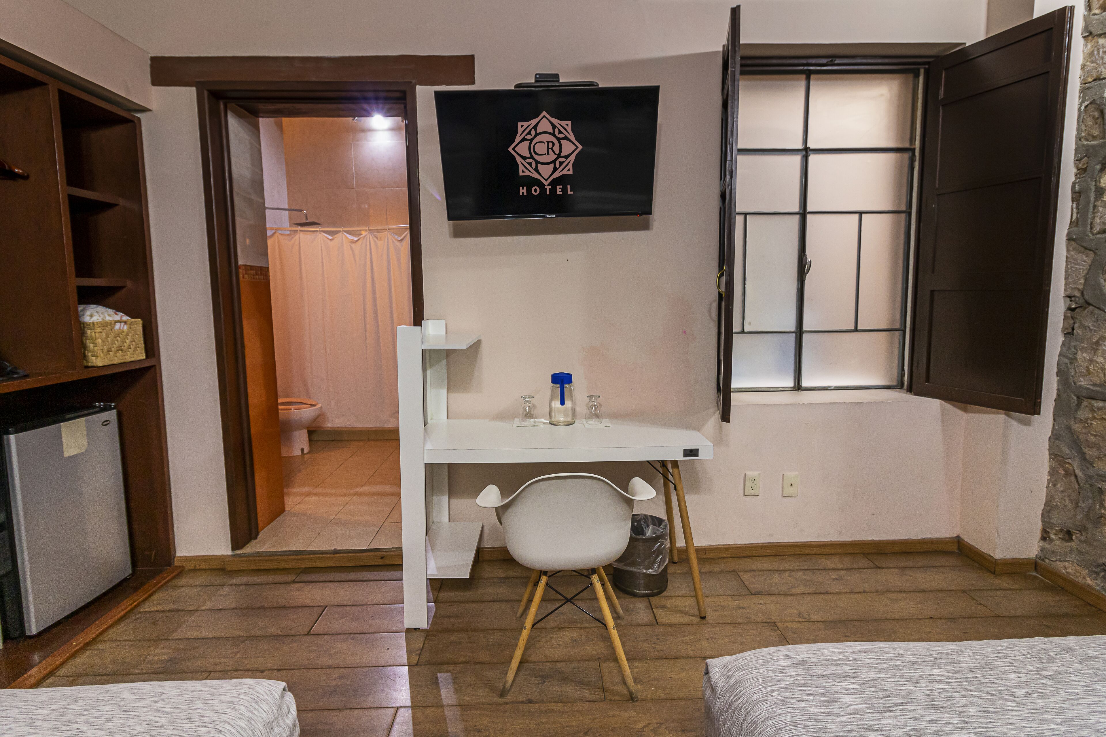 A compact hotel room with a small desk, a chair, a television, and a window with shutters.