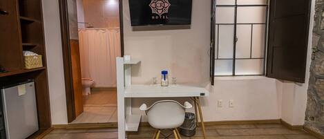 A compact hotel room with a small desk, a chair, a television, and a window with shutters.