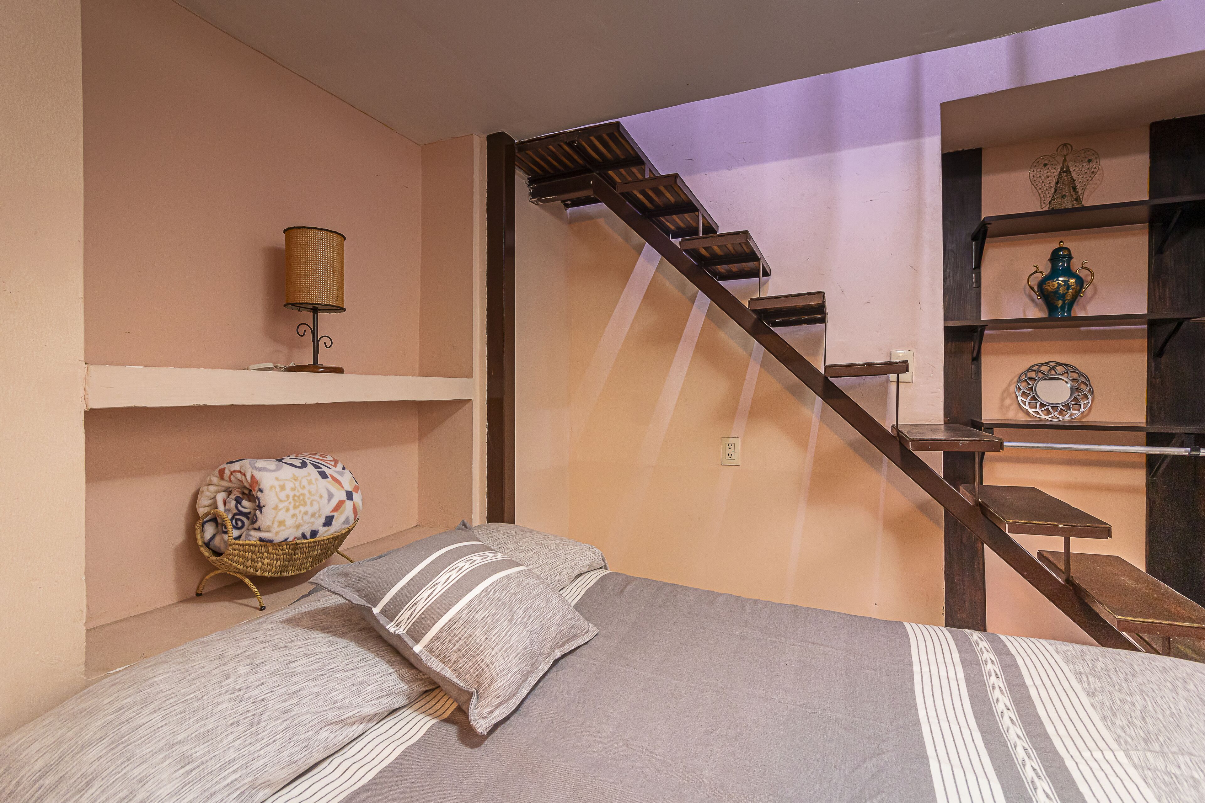 A bedroom with a bed, a lamp, a wicker chair, a staircase, and shelves with decorative items.