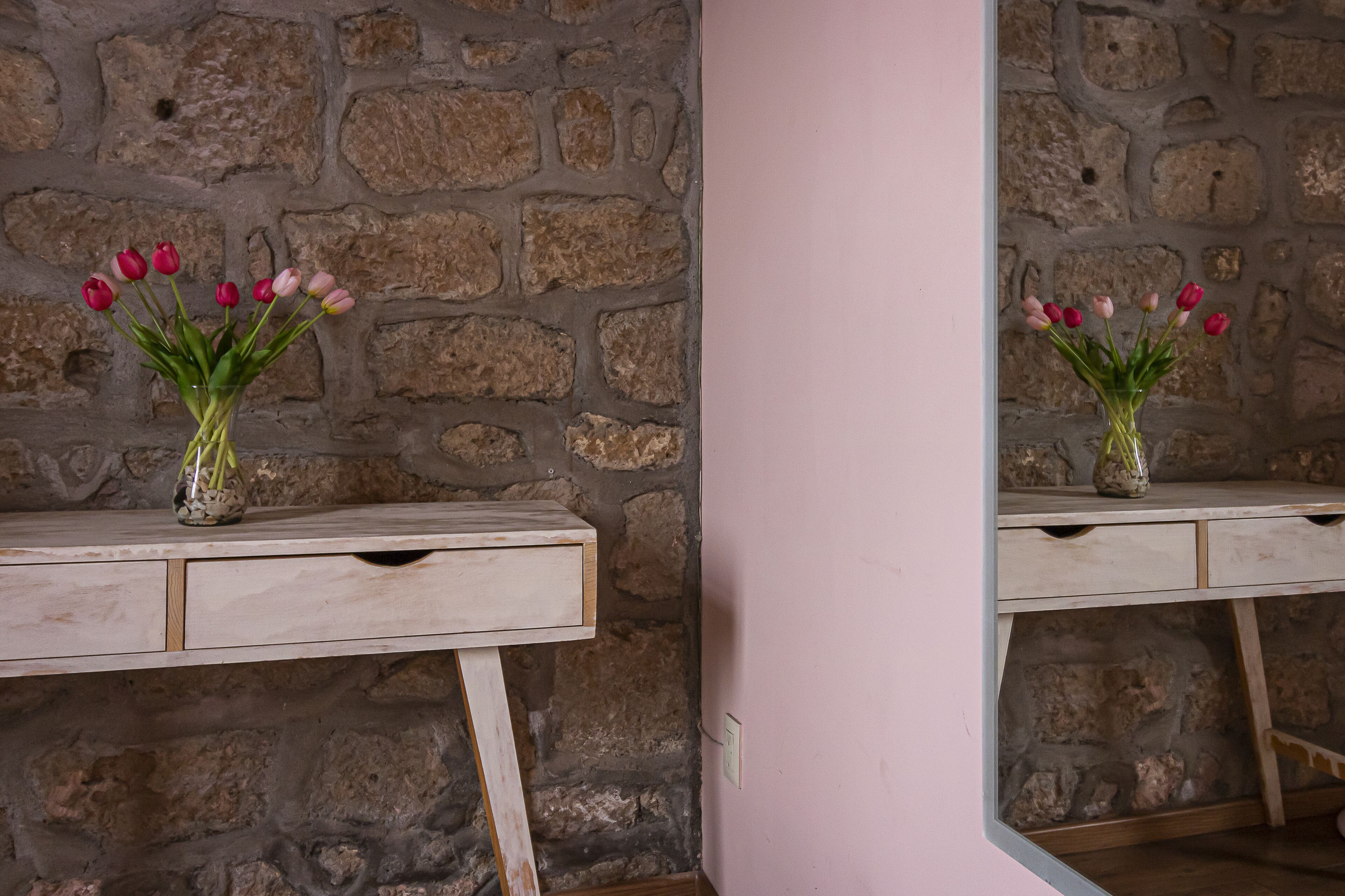 A stone wall with a mirror reflecting a vase of tulips on a wooden dresser.