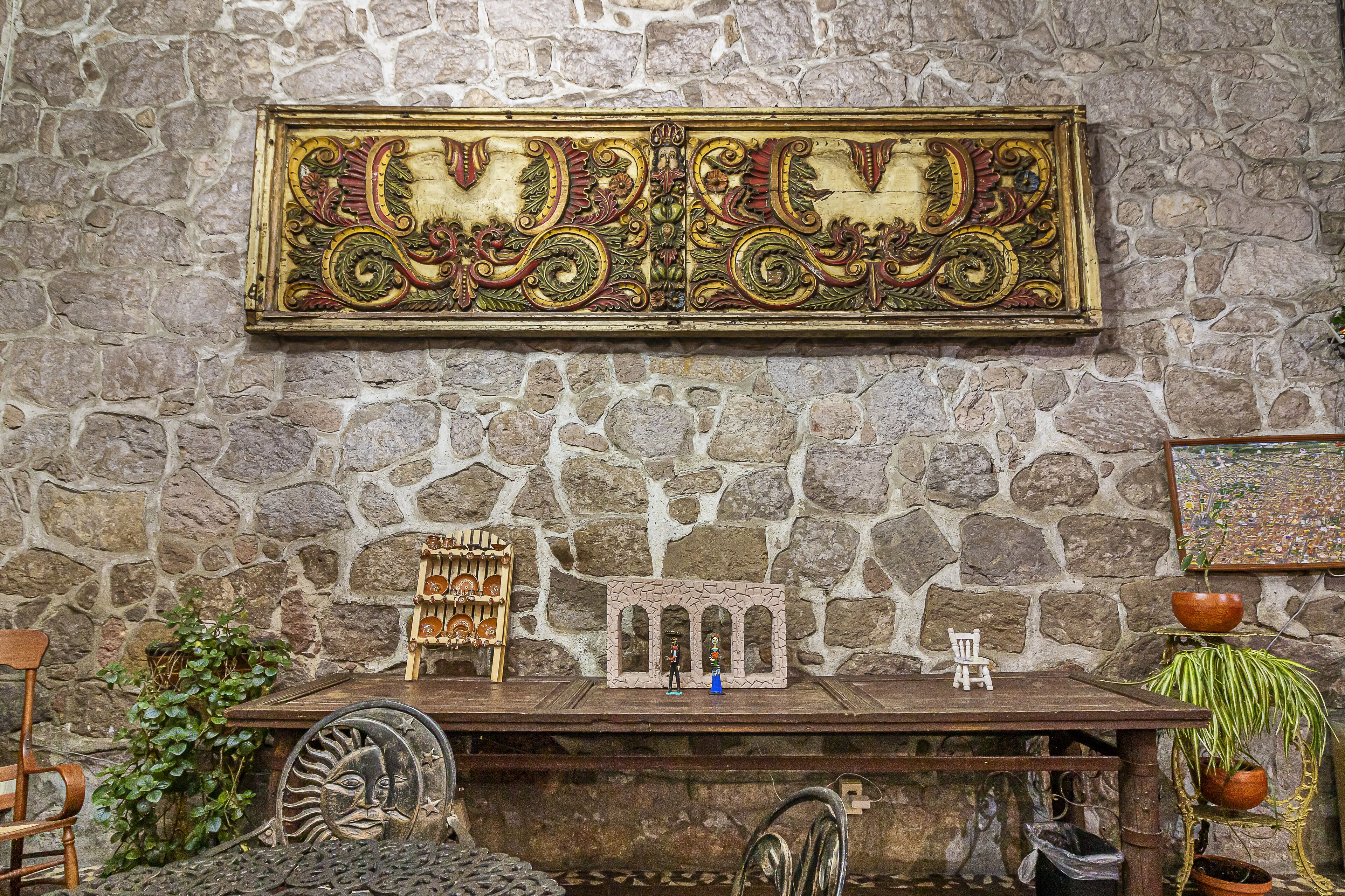 Un muro de piedra con un panel decorativo, una mesita con figuritas y una planta en maceta.