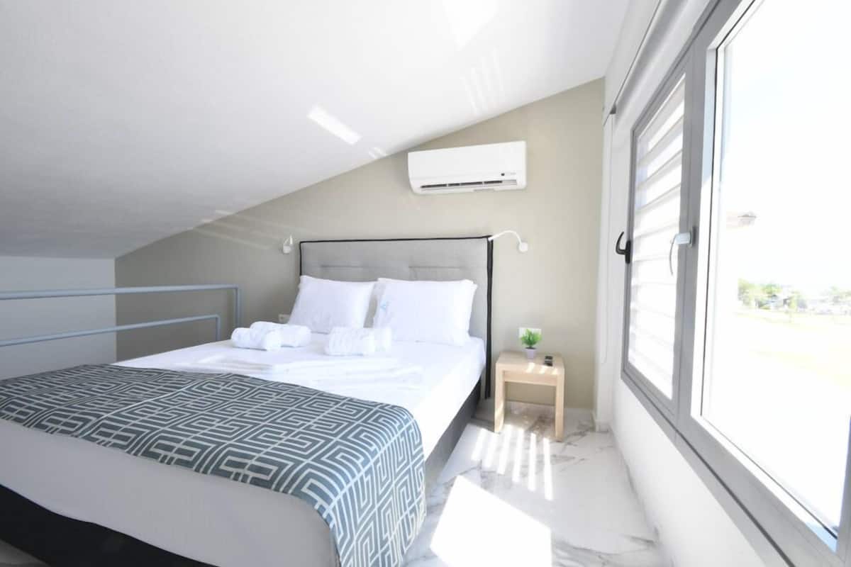 A bedroom with a bed, a bedside table with a plant, and a window with blinds.