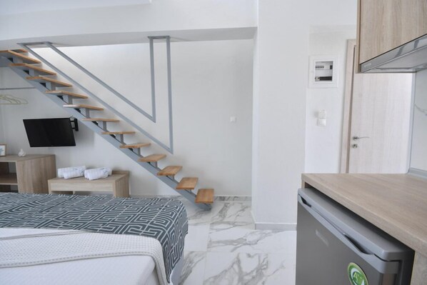 A bedroom with a bed, a TV mounted on the wall, a wooden staircase, a small wooden table with towels, and a compact kitchen area with a refrigerator.
