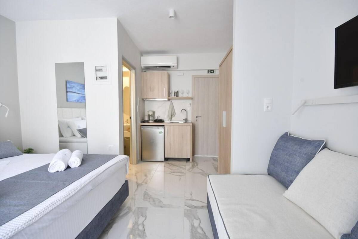 A modern hotel room with a bed, a sofa, a small kitchen area, and a TV mounted on the wall.