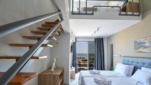 A hotel room with a bed, a desk, a chair, a small table, a plant, a painting, and a balcony with a view. - Alos Utopia (Volvi)