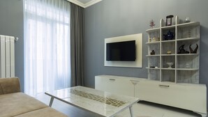 Un salon moderne avec un canapé beige, une table basse en verre, un meuble de télévision blanc doté d’un écran plat et une étagère en marbre agrémentée d’objets décoratifs.