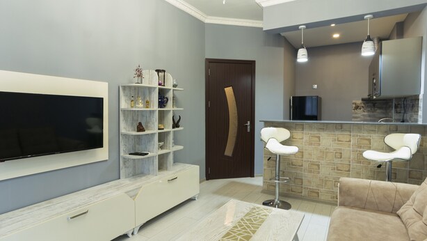 Un salon moderne doté d’un téléviseur à écran plat, d’un meuble de rangement blanc, d’un canapé beige et d’un coin cuisine avec un bar et des luminaires suspendus.