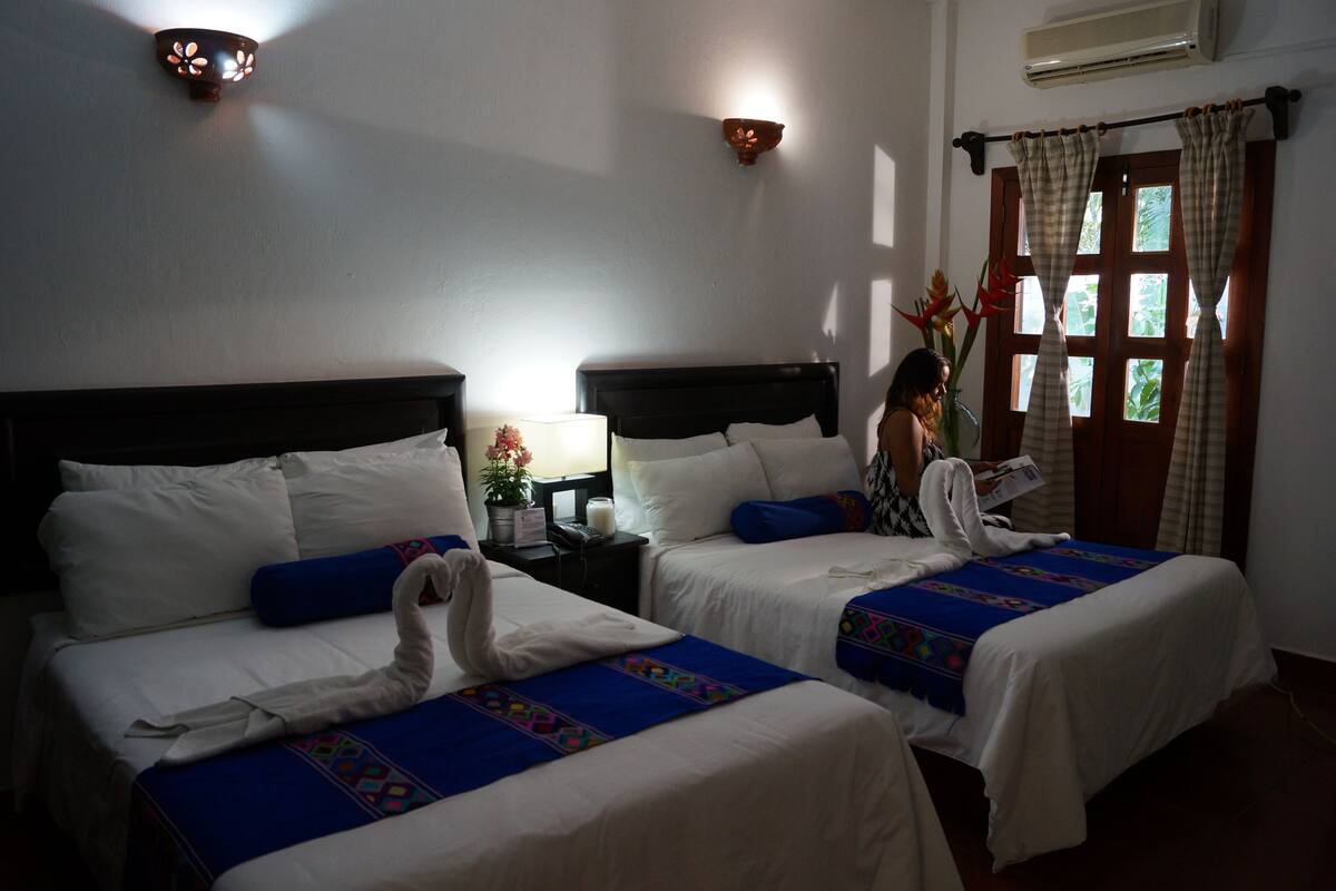 A hotel room with two beds, a nightstand with a lamp, and a window with curtains.