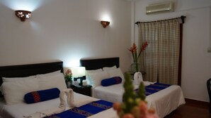 Ein Hotelzimmer mit zwei Betten, beide mit weißen Bettwäsche und blauen Zierkissen. Ein Nachttisch mit Lampe und eine Vase mit Blumen sind ebenfalls vorhanden.