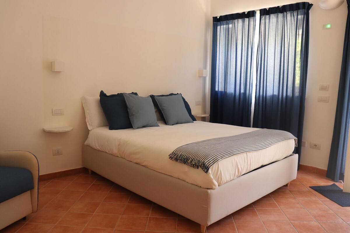 A bedroom with a bed, pillows, a bedside table, a chair, and a window with curtains.