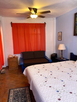 A bedroom with a bed, a nightstand, a lamp, a ceiling fan, and a window with red curtains. - Quinto Sol 2 Departamento (San Miguel de Allende)