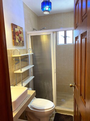 A bathroom with a toilet, a shower stall, a sink with a towel rack, and a decorative light fixture. - Quinto Sol 2 Departamento (San Miguel de Allende)