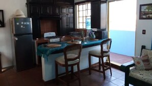 A kitchen with dark cabinets, a refrigerator, a stove, and a sink. There are two wooden chairs at the island. - Quinto Sol 4 Departamento (San Miguel de Allende)