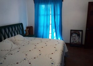 A bedroom with a bed, a window with blue curtains, a framed picture on the wall, and a wooden headboard.