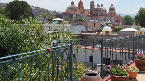 Una terraza en la azotea con barandilla de metal, plantas en macetas y vistas de un edificio histórico con cúpulas y campanarios.