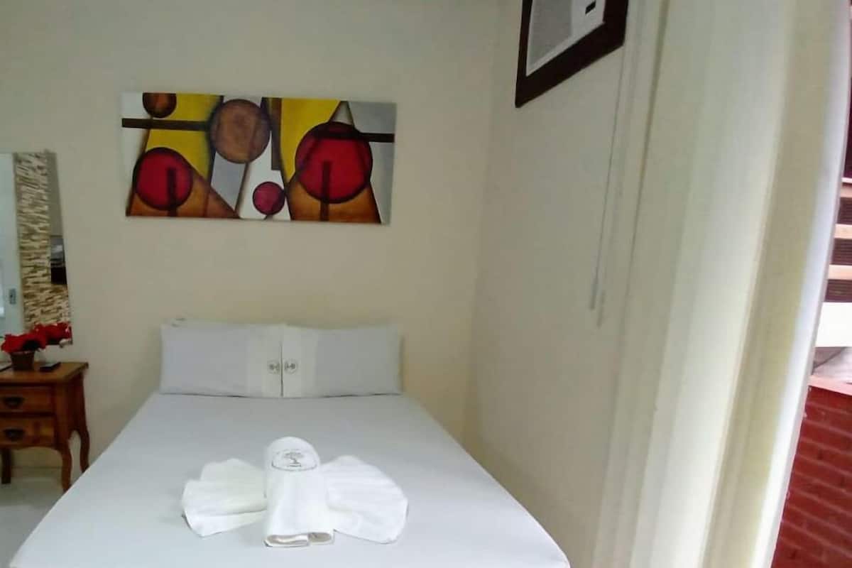 A neatly made bed with a quilted mattress and pillows, a painting on the wall, and a wooden nightstand with a vase of flowers.
