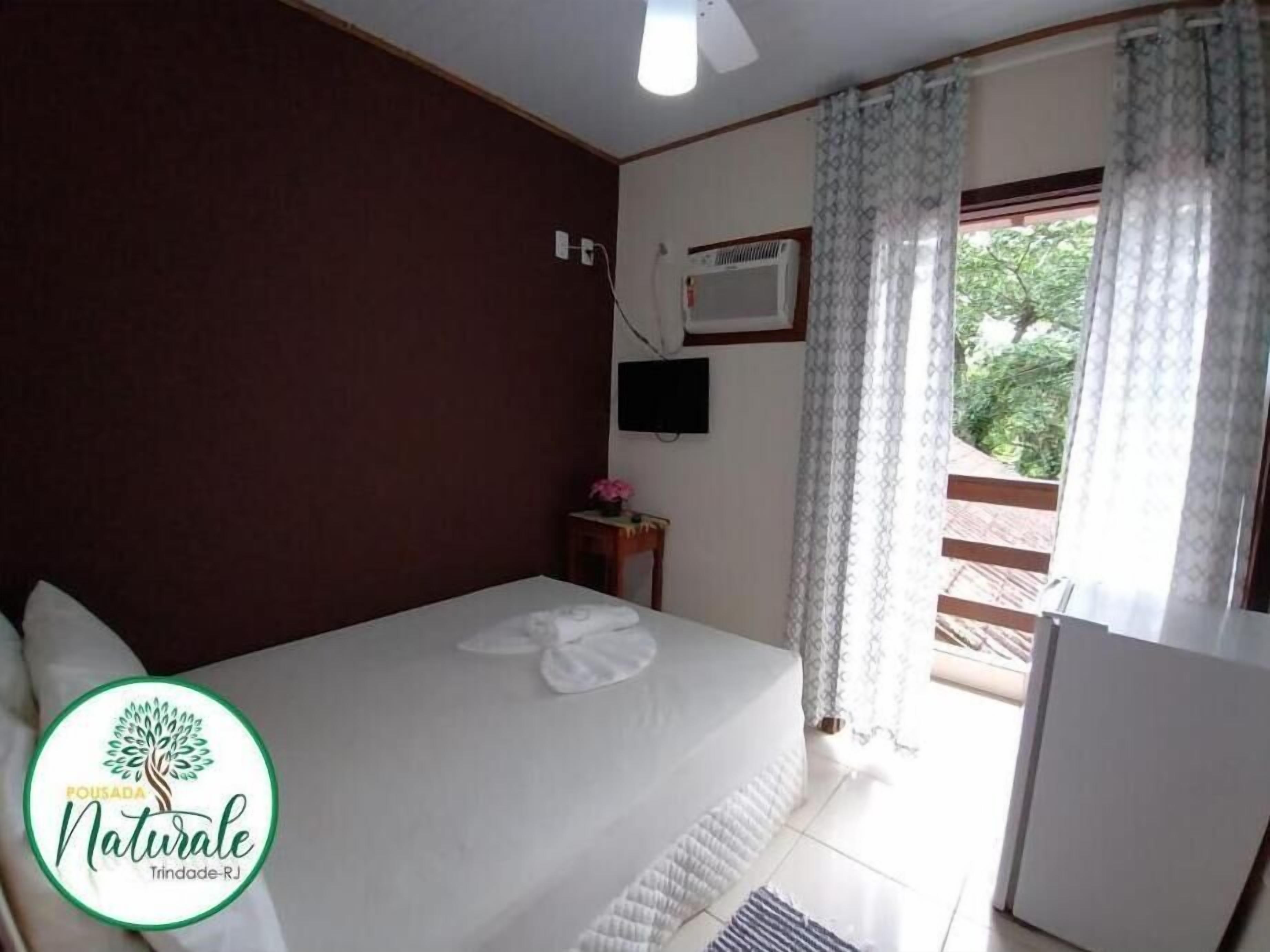 Quarto com cama, uma mesinha com uma flor, televisão e janela com cortinas.