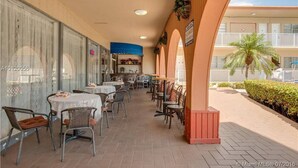 Un espace extérieur aménagé pour les repas, avec des tables et des chaises, une partie couverte avec un auvent bleu, et une vue sur un immeuble doté de balcons.