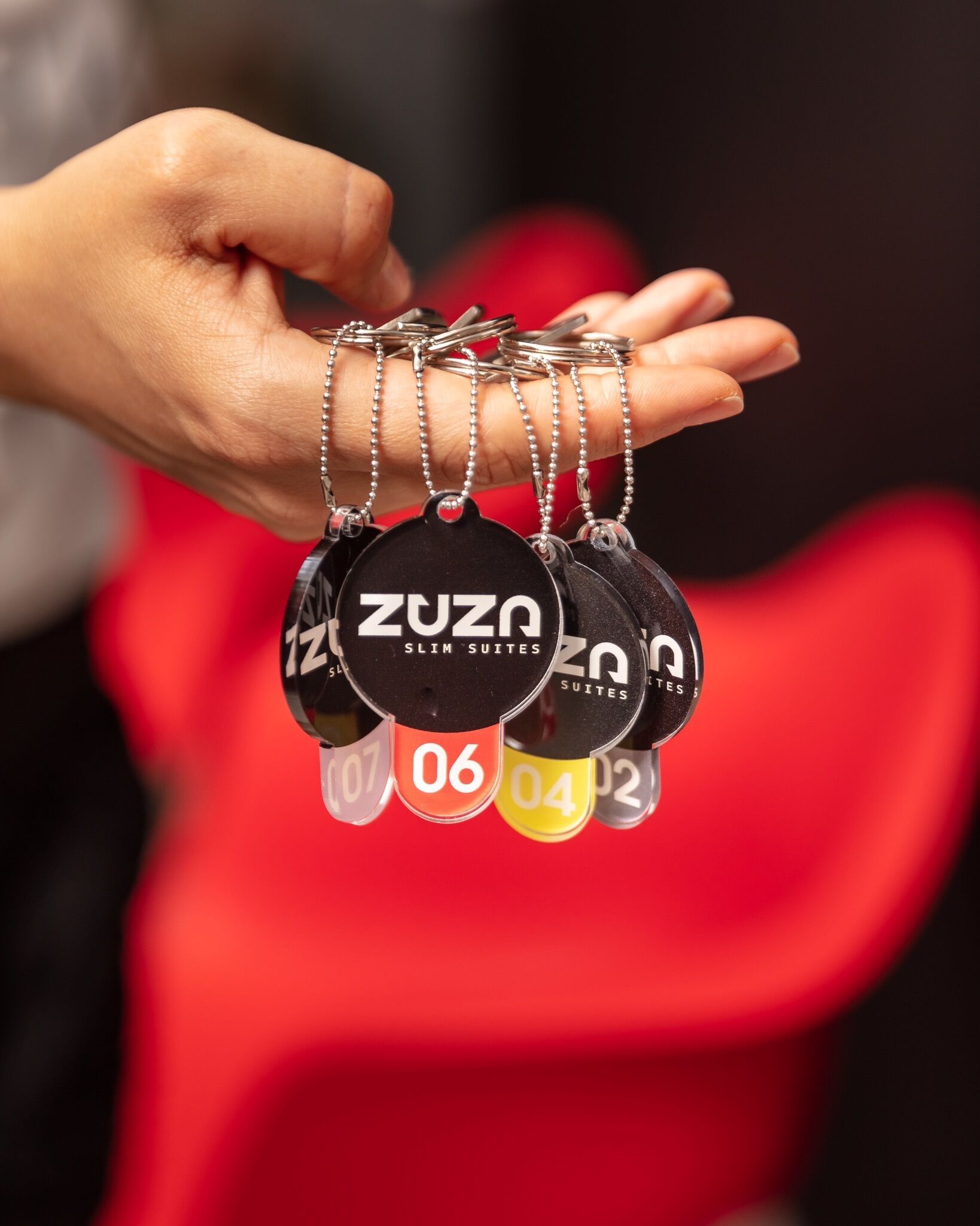 A hand holding a set of keychains with the text "ZUZA SLIM SUITES" and numbered tags.