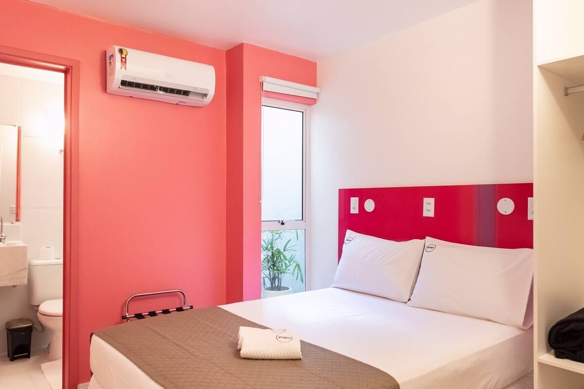 A hotel room with a bed, a window, an air conditioning unit, a small table, and a chair.