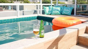 A swimming pool with a glass of beer and a bottle of Heineken, surrounded by green cushions with the ZUZO logo. - Zuza Slim Suítes (Maceió)