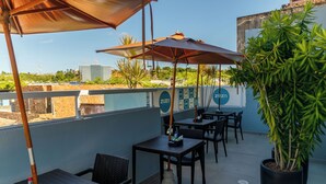 An outdoor patio with tables and umbrellas, a potted plant, and a view of buildings in the background. - Zuza Slim Suítes (Maceió)