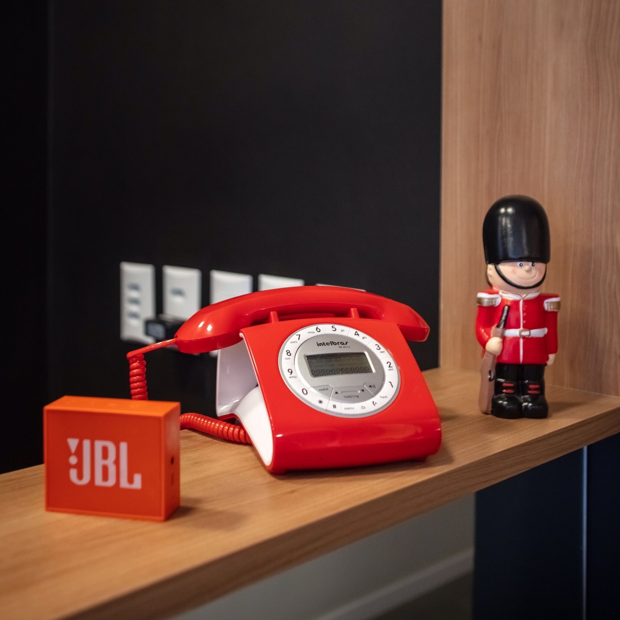 Un teléfono antiguo de disco rojo sobre una estantería de madera, con una pequeña figurina de un guardia y un cartel que dice "JBL".