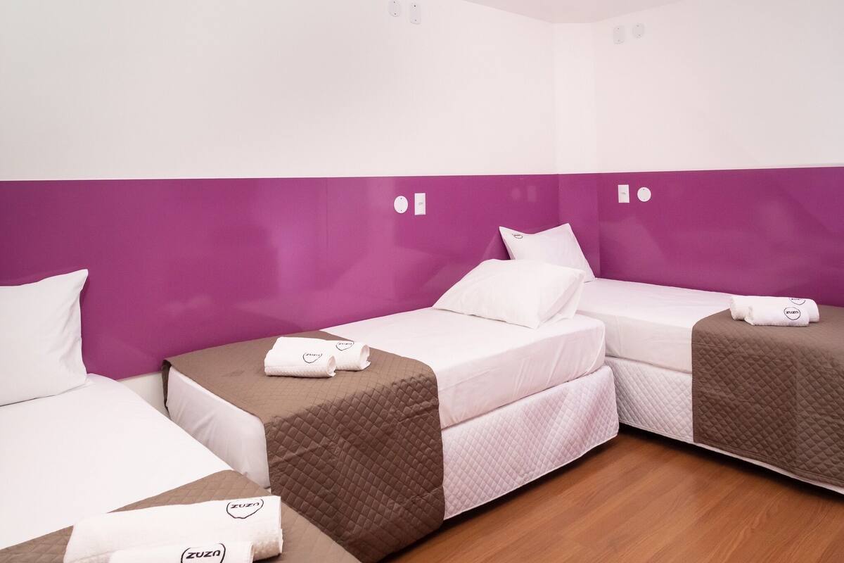 A hotel room with three beds, each with a white pillow and a brown quilted blanket. The room has purple walls and wooden flooring.