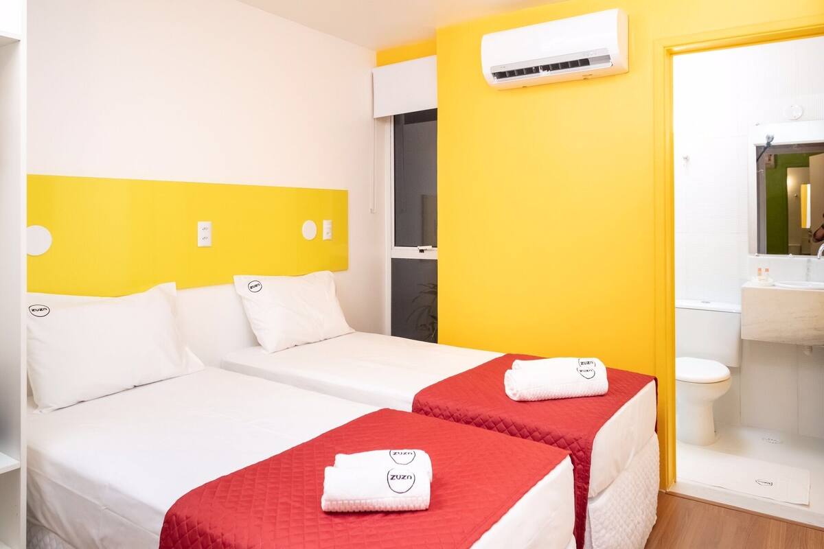 A hotel room with two beds, each with a red quilted bedspread and white pillows, and a yellow accent wall.