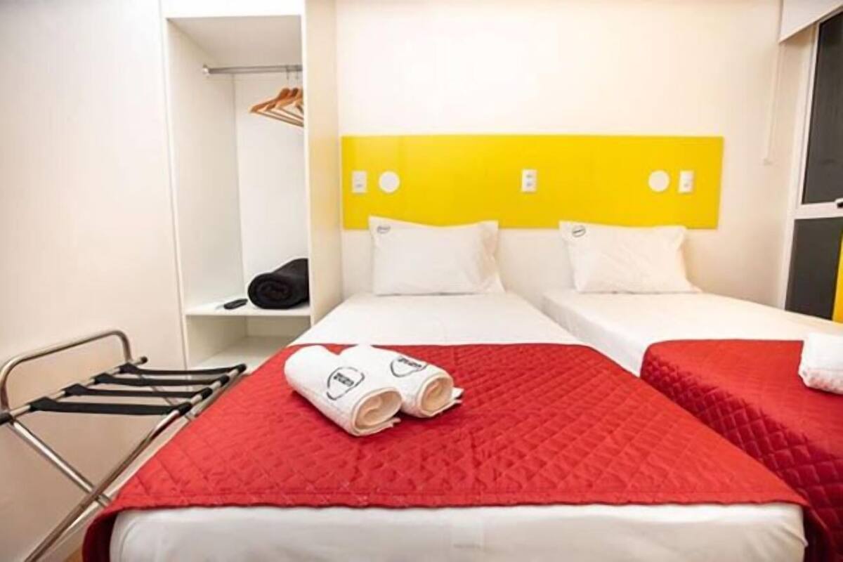 A hotel room with two beds, a red quilted bedspread, a white headboard, and a metal rack with a black bag.